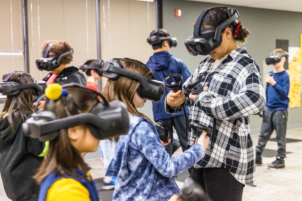 A group of children in VR headsets play together while an adult helps one of them with a controller.