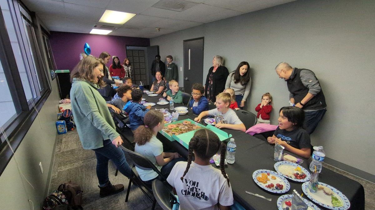 Kids eat pizza around a long table at a birthday party while parents look on.