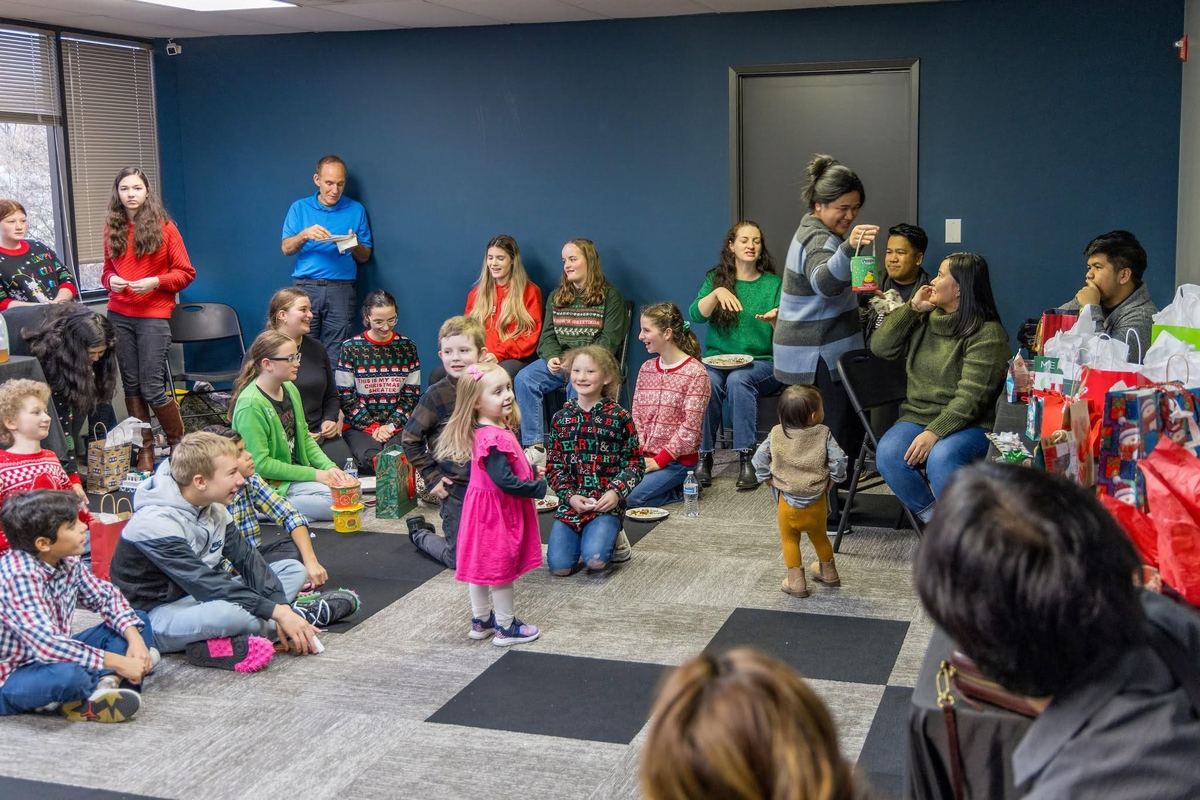 Kids sit on the floor and families fill the room at a holiday party, with presents piled to one side.