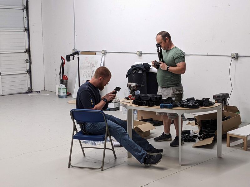 Two people unbox and set up VR blasters and headsets on a table in the warehouse.