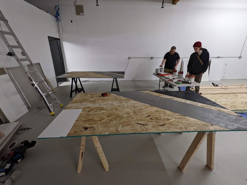 Two people build plywood panels on sawhorses in an empty white warehouse.